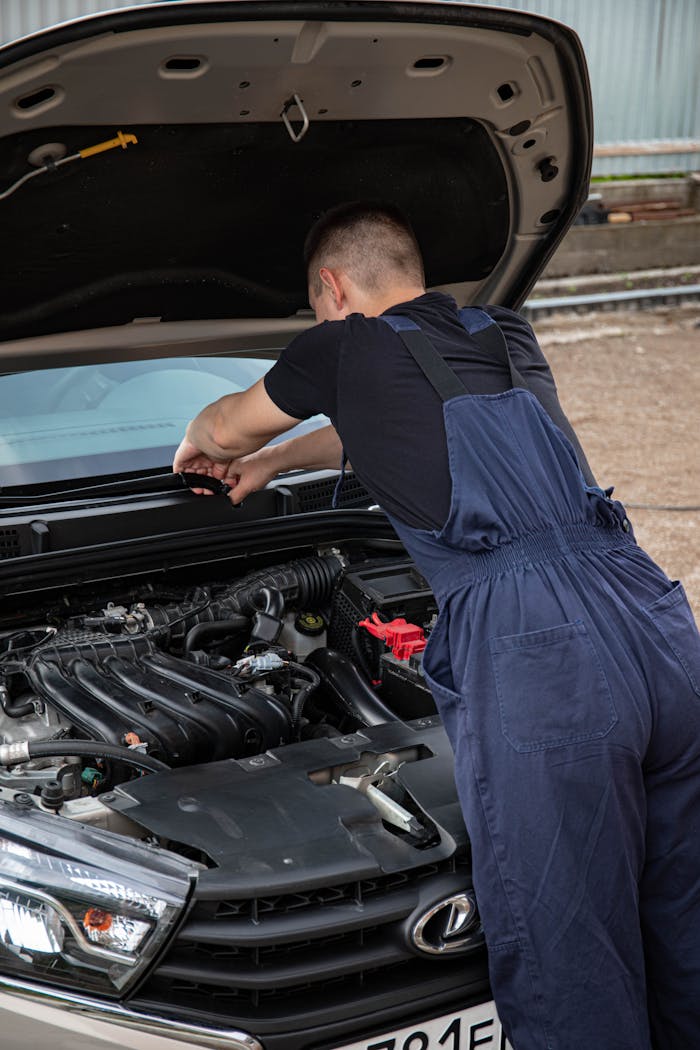 Mechanic in blue overalls repairing a car engine outside during the day. Professional auto maintenance.