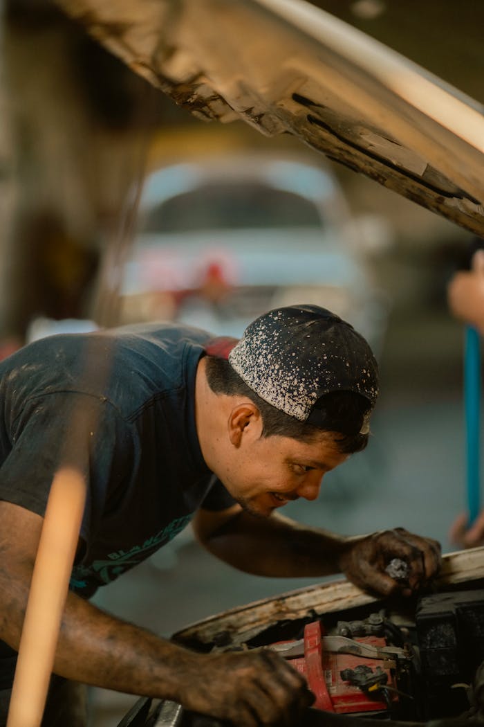 Mechanic repairing car under open hood, focused, wearing cap, selective focus.